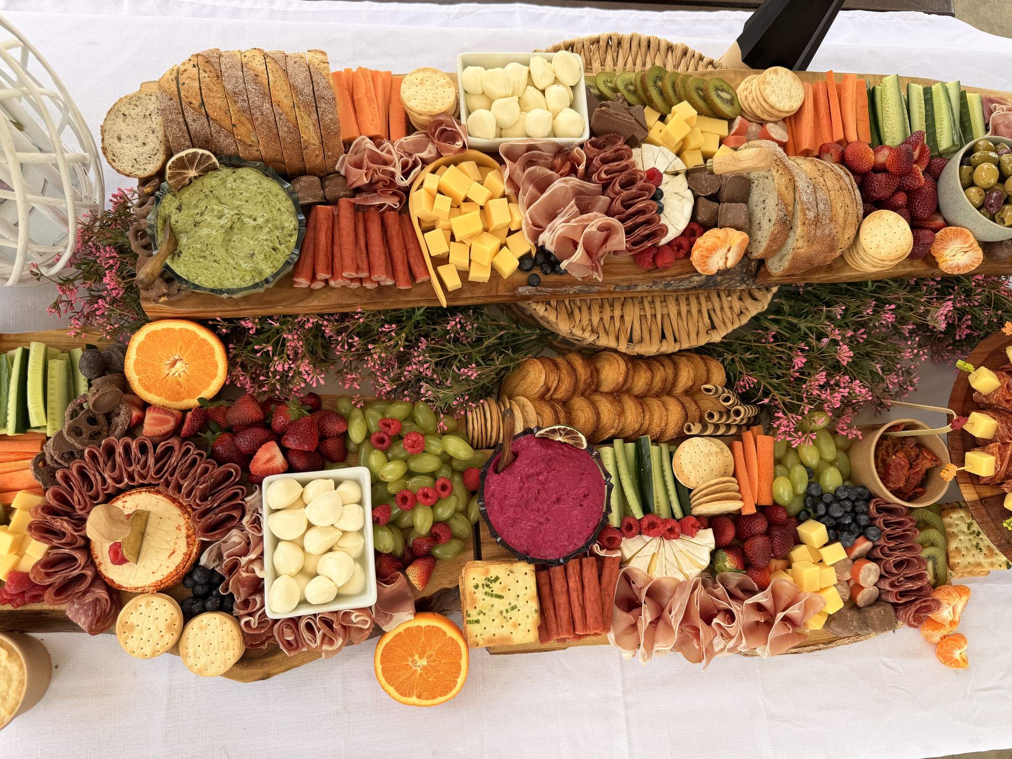 A long grazing table from above, filled with cheeses, cured meats, fruit, dips and pink flowers